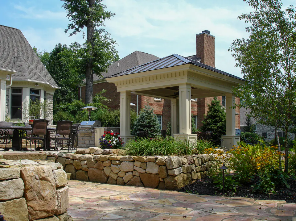 Covered pavilion with painted posts, standing seam roof, stone wall edge, paver patio, lawn, and layered planting