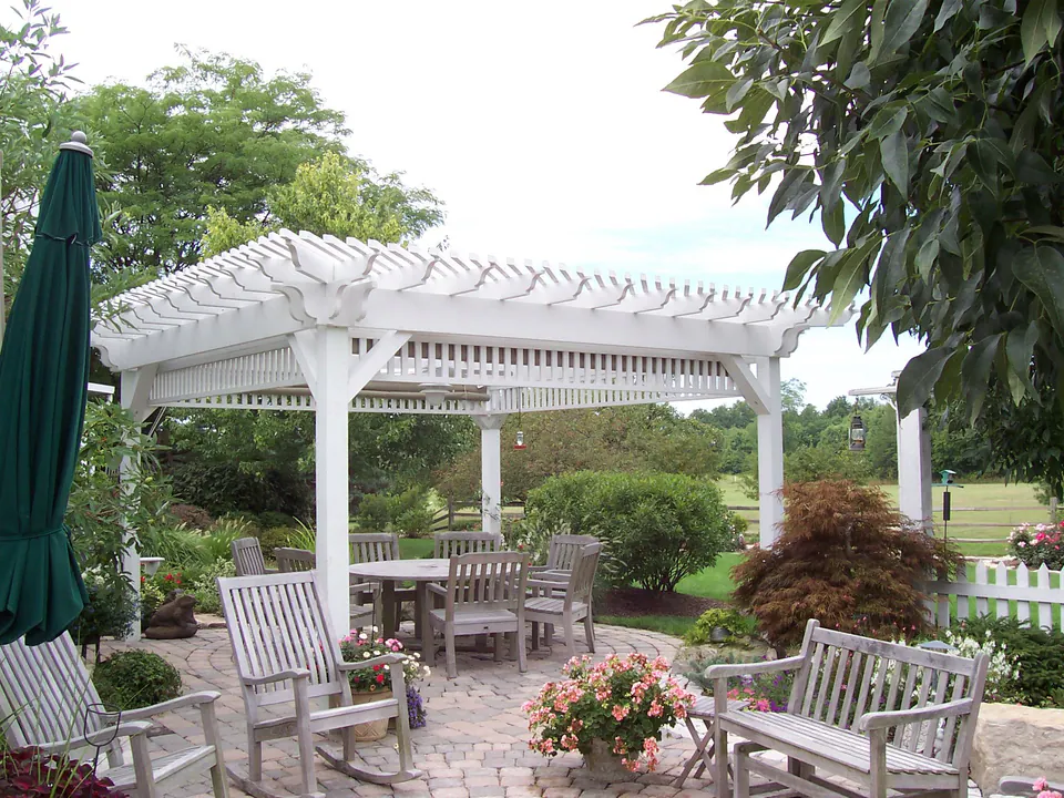 White pergola over a paver patio with dining table, rocking chairs and container flowers