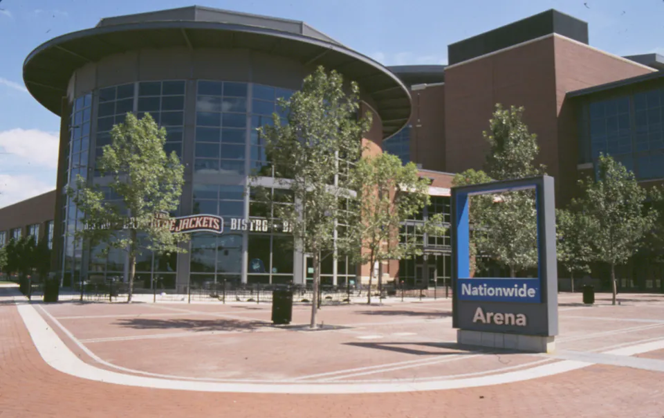 Nationwide Arena sign on a brick paver plaza with young street trees in front of the glass-fronted arena
