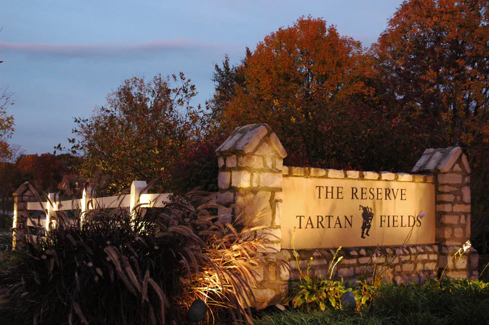 Lit stone entry monument reading The Reserve at Tartan Fields at dusk, with ornamental grasses and fall trees