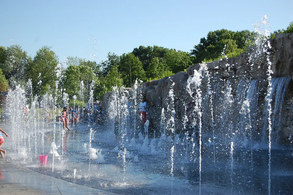 Splash pad with vertical water jets in front of a stone waterfall wall, with children playing
