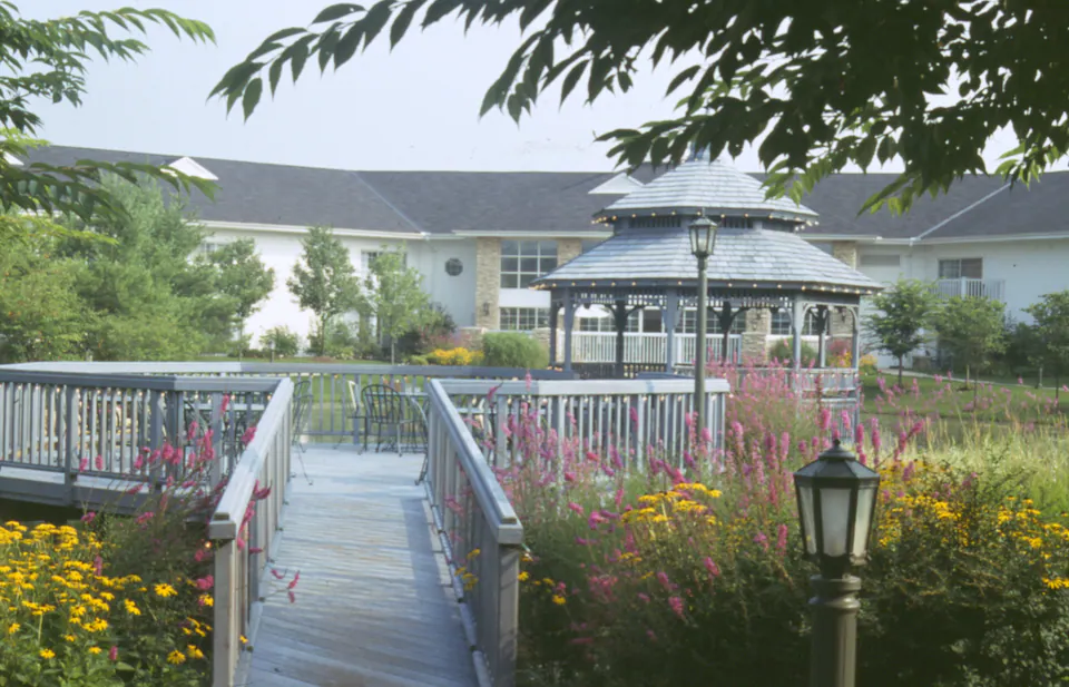 Wood footbridge and railed walk leading to a gazebo beside a pond, with wildflowers and a lamp post at a community clubhouse