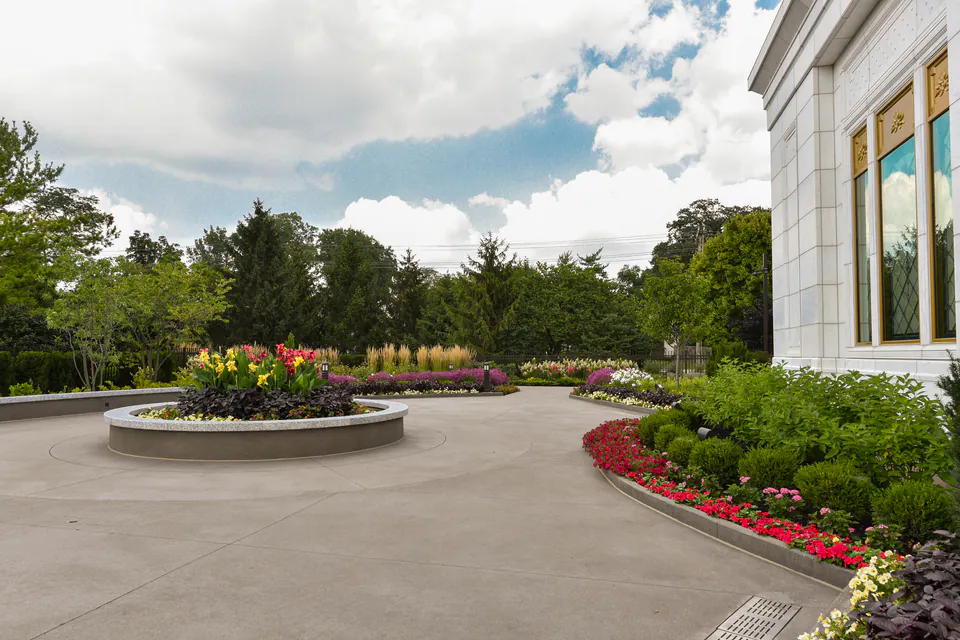 Concrete plaza with a round raised planter of annuals and curved beds of red annuals beside a white stone building