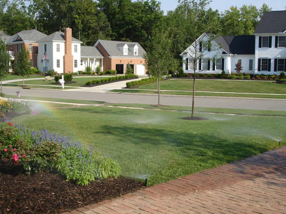 Sprinklers spraying over a community lawn and planting bed beside a brick walk, with townhomes behind