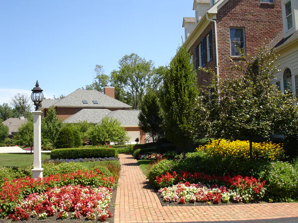 Brick walk between beds of red and white annuals, a lamp post, and clipped shrubs beside a brick house