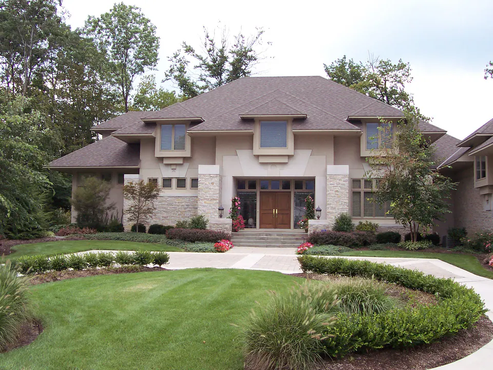 Prairie-style stone house with a circular lawn, hedges, and a mulched bed of shrubs in the foreground