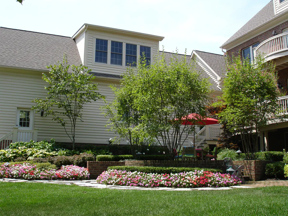 White house with a red umbrella, curved brick seat walls, and beds of pink and white annuals beside a lawn