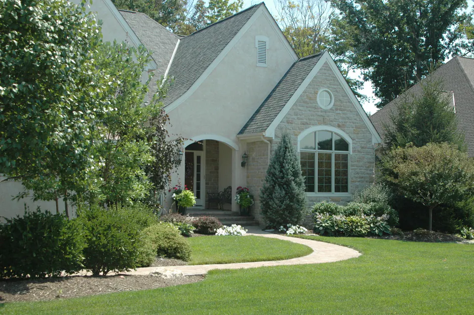 Stucco house with an arched entry, a curved concrete walk, white annuals, and clipped foundation shrubs