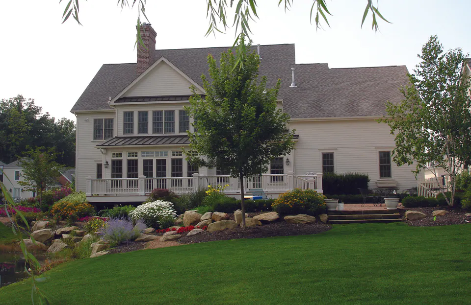 Cream house with a wide porch above a boulder-edged planting bed and a maintained lawn under a willow