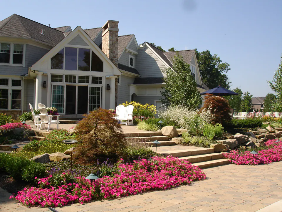 House with a stone terrace and steps above beds of pink annuals, a Japanese maple, and a paver drive