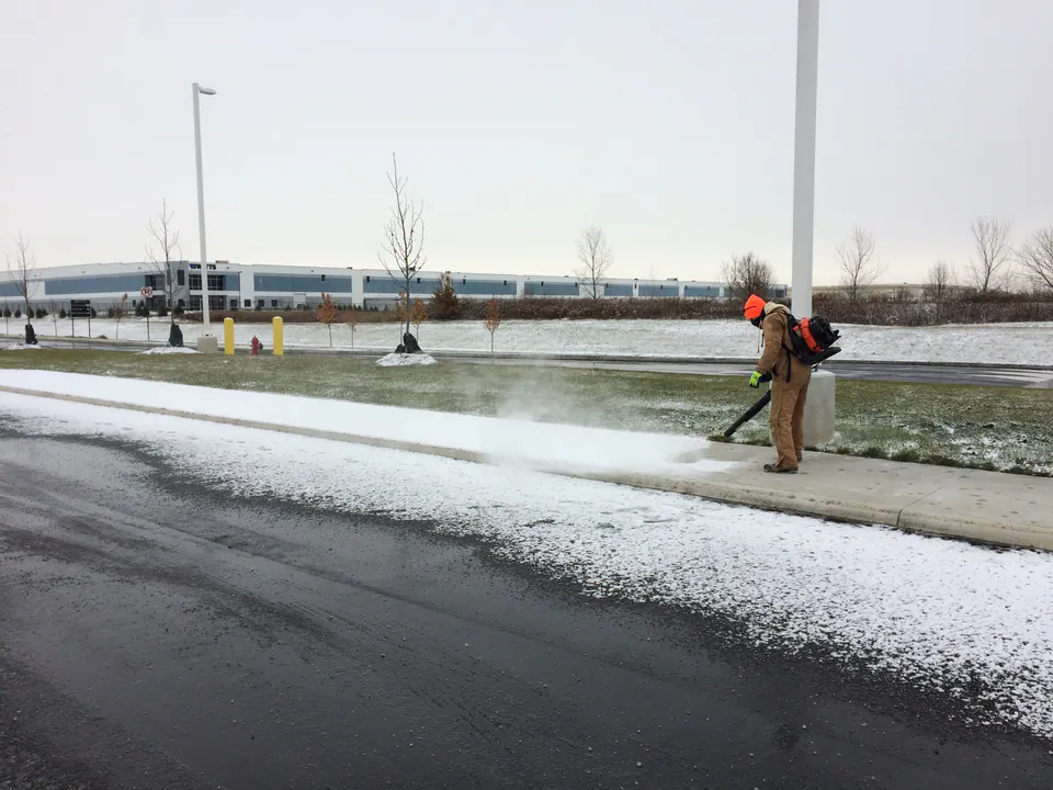 Crew member with a backpack blower clearing light snow from a sidewalk in front of a distribution center