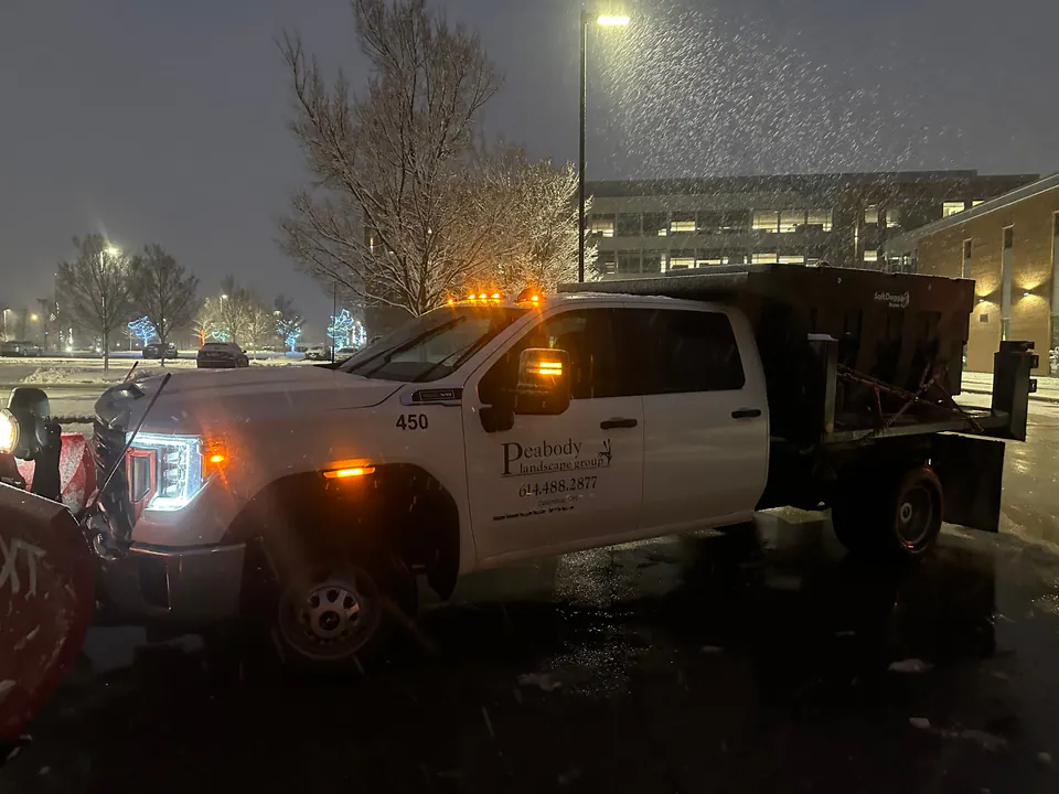 Peabody Landscape Group plow truck with a salt spreader in falling snow outside an office building at night